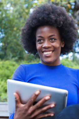 Parkta bankta oturan ve yeni başlatılan projede çalışırken tablet kullanan içerikli Afrikalı Amerikalı kadın serbest yazar