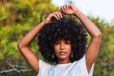 Çekici, duygusuz, kıvırcık saçlı, kollarını kaldırmış, doğadaki yeşil ağaçların arka planındaki kameraya bakan Afro-Amerikan kadın.