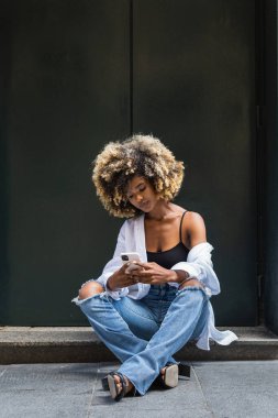 Çapraz bacaklı ve cep telefonuyla oturan yırtık kot ve beyaz gömlekli çekici Afro-Amerikan kadın.