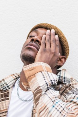 Şık, kareli gömlekli, yorgun bir Afro-Amerikan adam. Gözü kapalı gri duvara sürtünüyor.