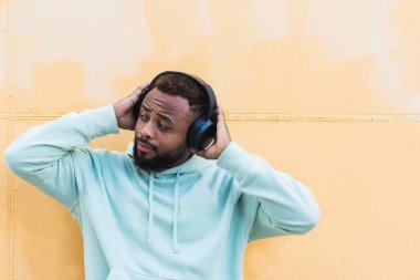 Yetişkin sakallı, mavi kapüşonlu Amerikalı erkek. Sarı arka planda kablosuz kulaklıklı müzik dinliyor.