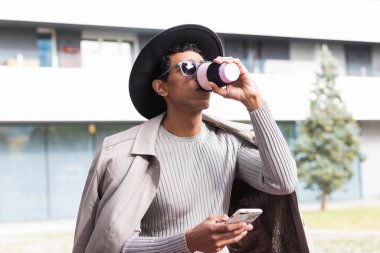 Yetişkin, modaya uygun giyinmiş, güneş gözlüklü, cep telefonuyla sohbet eden ve sıcak içeceklerle şehirde dinlenen yetişkin erkeklerin yan görüntüsü.