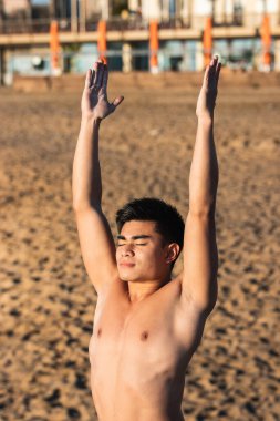 Kırpılmış gömleksiz genç Asyalı erkek. Kollarını kaldırıp plajda gözleri kapalı yoga yaparken poz veriyor.