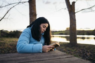 Kitap okuyan bir kadın kışın gün batımında güzel bir gölün kenarında bir masada oturuyor.