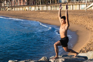 Tüm vücudu çıplak genç Asyalı bir erkeğin kumsalda yoga yaparken kollarını kaldırarak duruşu.