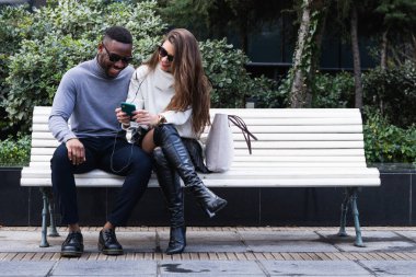 Parkta birlikte vakit geçirirken bankta oturan ve cep telefonlarını karıştıran çok ırklı mutlu çiftin tüm vücudu.