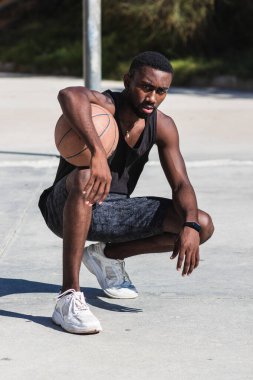 Azimli Afro-Amerikan erkek spor giyim şehirde basketbol sahasında top ile çömelmiş ve kameraya bakıyordu.