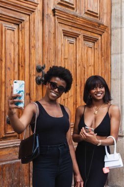 Sokakta yan yana dururken cep telefonlarıyla selfie çeken Afrikalı Amerikalı kız kardeşler.