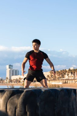 Kendine güvenen genç atletik Asyalı erkek şehir seti üzerinde araba lastiğiyle antrenmana hazırlanırken enerjik atlama egzersizi yapıyor.