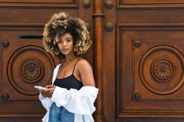 Çekici Afro-Amerikan kadın şık giyinmiş girişte duruyor ve cep telefonuyla sörf yapıyor.