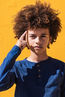 Afro kıvırcık saçlı düşünceli genç çocuk tapınağa dokunuyor ve sarı arka planda kameraya dikkatlice bakıyor.