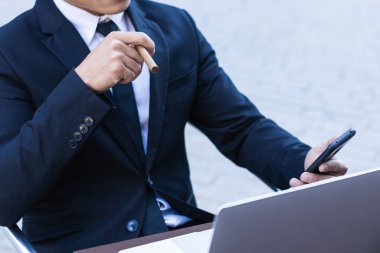 Kendine güvenen Asyalı erkek girişimci kafeteryada masada oturmuş, akıllı telefonlara bakarken puro içiyor.