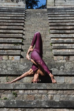 Spor giyim sektöründe ince bir kadın Asya 'da yoga yapıyor ve taş merdivenlerde baş aşağı dengede duruyor.
