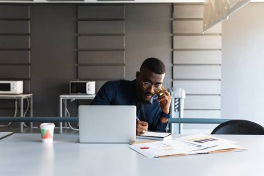 Gözlüklü Afro-Amerikan erkek ve masada resmi kıyafetler cep telefonuyla konuşuyor ve not defterine, netbook 'un yanındaki notlara ve çalışma alanındaki açık alan içecekleriyle birlikte dokümanlara not alıyor.