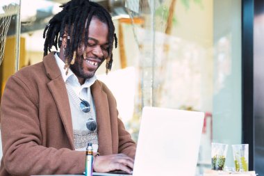 Odaklanmış neşeli hippi, siyah erkek, dağınık saçlı, laptopla masada oturuyor ve açık hava restoranında uzak bir proje üzerinde çalışıyor.
