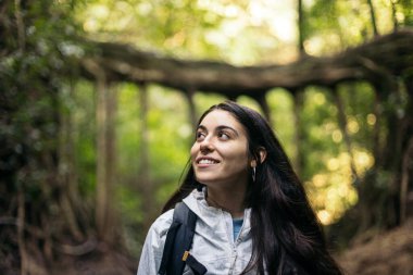 Yağmurluk ve sırt çantalı genç bir kadın büyük ağaçlarla dolu bir ormanda yürüyor..