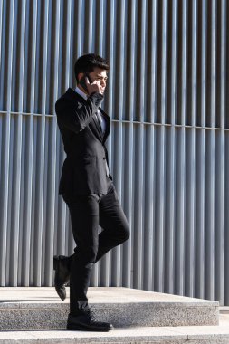 Siyah takım elbiseli neşeli erkek girişimci modern binanın yanındaki merdivenlerde duruyor ve cep telefonuyla iş konularını tartışıyor.