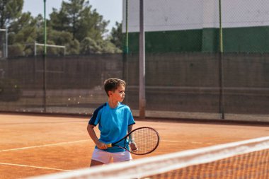 Spor giyim kuşağında tenis oynayan küçük çocuk tenis kortunda elinden gelenin en iyisini yapmaya çalışıyor.