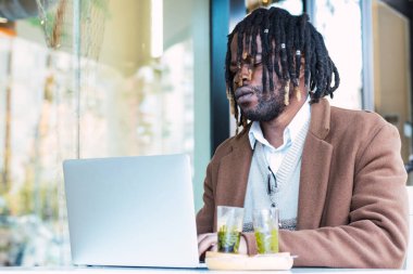 Ciddi odaklı hippi siyahi erkek, Afro saçlı, dışarıda bir restoranda uzaktan kumandalı bir proje üzerinde çalışırken dizüstü bilgisayarla masada oturuyor.