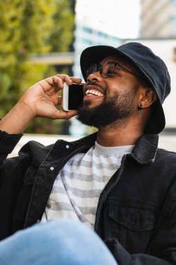 Parkta dinlenirken ahşap bankta oturan ve cep telefonuyla konuşan, güler yüzlü Afro-Amerikan erkek.