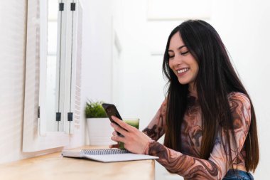 Tezgahta oturmuş gülümseyen bir kadın barda yeşil smoothie ile akıllı telefondan bakarken not defterinde yazıyor.