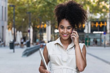 Afro saç stili pozitif Afrikalı kadın cep telefonuyla konuşurken karnına dokunuyor modern ofis binasının yakınında bir laptop taşıyor.