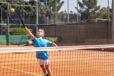 Spor giyim kuşağında tenis oynayan küçük çocuk tenis kortunda elinden gelenin en iyisini yapmaya çalışıyor.