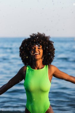 Mayo giymiş neşeli Afro-Amerikalı kadın ve dalgalı saçlarıyla denizde durup yaz tatilinde eğlenirken su sıçratıyor.