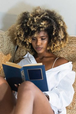 Çekici Afro-Amerikan kadın kıvırcık saçlı, dağınık yatakta oturuyor ve sabahları kitap okuyor.