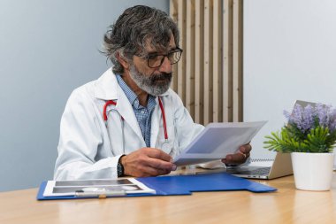 Tıp bürosunda masada oturan ve klinikte çalışırken röntgen çeken yaşlı bir erkek doktor.