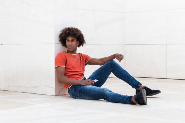 Modern binanın fayanslı zemininde otururken elinde cep telefonu tutan, günlük kıyafetleriyle kıvırcık saçlı, ciddi Afro-Amerikan genç adamın yan görüntüsü.