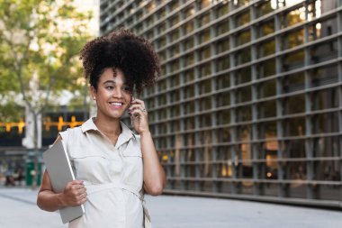 Afro saç stili pozitif Afrikalı kadın cep telefonuyla konuşurken karnına dokunuyor modern ofis binasının yakınında bir laptop taşıyor.