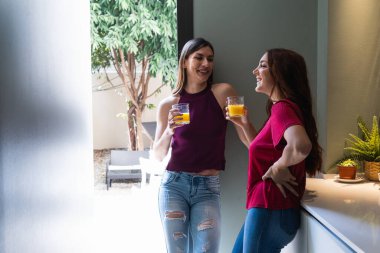 Çekici genç bayan arkadaşlar gündelik giysiler giyip taze portakal suyunun tadını çıkarıyor ve modern aydınlık mutfakta dururken hoş sohbetler ediyorlar.