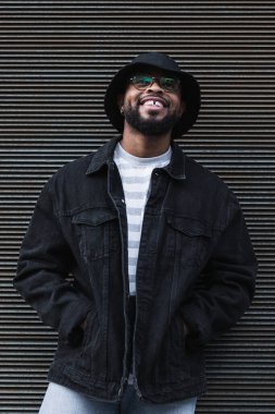 Gözlüklü, gözlüklü, modaya uygun kıyafet ve şapka takan parlak bir şekilde gülümseyen ve kepenkli duvara bakarken yüzünü çeviren mutlu Afro-Amerikan sakallı adam.