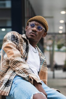 Moda desenli tişörtlü genç siyahi adam modern şehrin sokaklarında serinlerken kaşlarını çatıyor ve yüzünü çeviriyor.