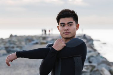 Spor kıyafetleri içinde genç bir Asyalı erkek dışarıda spor yaparken bulanık deniz ve günbatımı gökyüzüne karşı dururken kollarını ve omuzlarını esnetiyor.