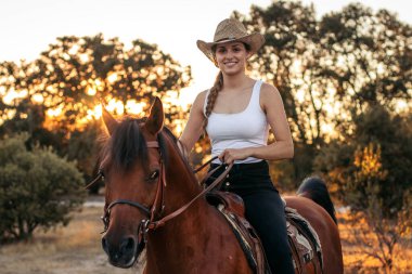 Güneş batarken kırsalda ata binen genç bir kadın. Görüntü, özgürlük ve doğa ile bağlantı hissini iletir.