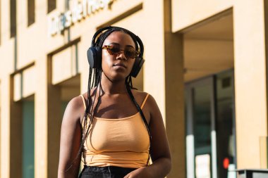 Çağdaş bina duvarına yaslanmış şık ceketli genç siyahi bir kadın ve şehir caddesinde kameraya bakarak kulaklıkla müzik dinliyor.