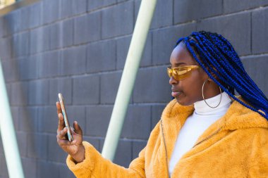 Mavi örgülü, sarı elbiseli, şık siyah bir kadın sokakta akıllı telefonlara bakıyor.