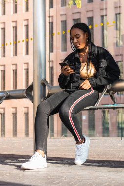 Neşeli siyah kadın, gündelik giysiler içinde, gülümseyen ve yüzünü çeviren modern otobüs durağında bankta otururken.