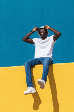 Parlak renkli çitlerde oturup güneşli yaz gününün tadını çıkarırken panama şapkalı ve güneş gözlüklü, mutlu gülümseyen, gündelik kıyafetli, iyimser genç bir erkek.