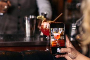 Tanımlanamayan bir kadının akıllı telefonuyla kokteyl çekerken yakın plan fotoğrafı. Bir barda garsonluk yapıyor.