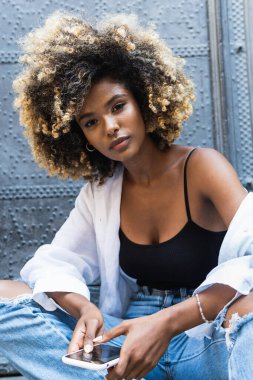 Serene çekici Afro-Amerikan kadın gündelik kıyafetleriyle oturmuş cep telefonuyla konuşuyor ve kameraya bakıp gülümsüyor.