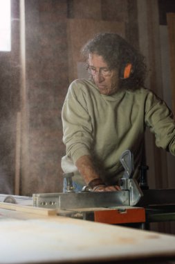 Korunaklı kulaklıklarla odaklanmış erkek katılımcı tozlu atölyede tahta testereyle tahta kesiyor.