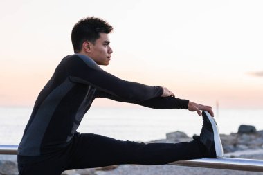 Spor kıyafetleri içinde genç Asyalı bir erkeğin gün batımında spor yaparken ısınırken metal korkulukların yanında bacaklarını esnetmesinin yan görüntüsü.