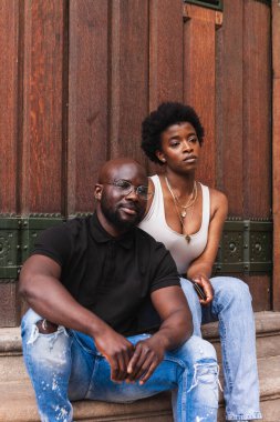 Ciddi genç Afro-Amerikan kadın ve erkek arkadaşı birlikte oturup ahşap duvara bakıyorlar.