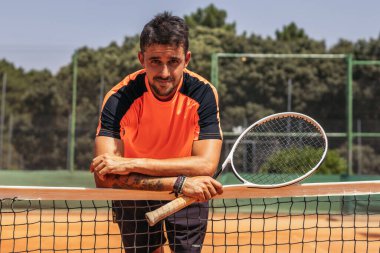 Spor giyimli bir adamın portresi, tenis kortunda tenis oynayan, internete yaslanan ve kameraya bakan.