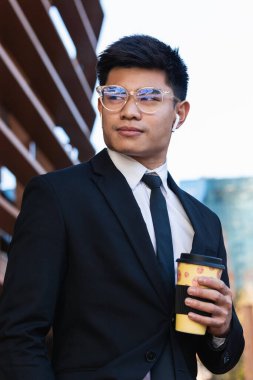 Resmi takım elbiseli, kablosuz kulaklıklı, kağıt bardak kahveyle caddede dikilen ve gözlerini kaçıran erkeksi Asyalı girişimci.