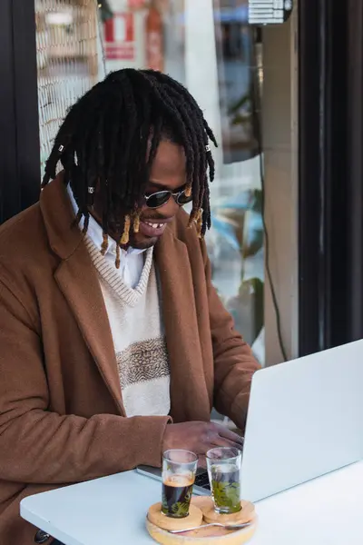 Stock image Side view of cheerful focused hipster black male with Afro in dreadlocks sitting at table with laptop while working on remote project in outdoor restaurant