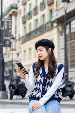 Asian young woman in fashionable clothes sitting on a bench in a European city waiting for someone while using the smartphone clipart